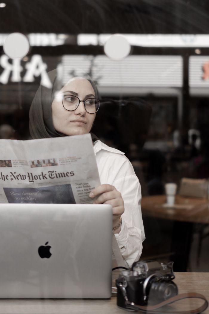 Services Woman in hijab reads a newspaper in a cafe, with a laptop and camera on the table.